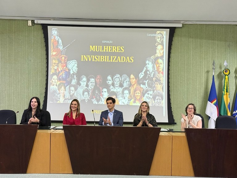 A foto mostra cinco integrantes em uma mesa de debates no auditório. Ao fundo, uma tela de projeção exibe o cartaz da exposição "Mulheres Invisibilizadas", acompanhado pelas bandeiras de Pernambuco e do Brasil no palco.