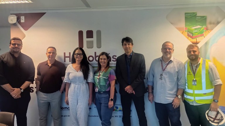 A imagem mostra um grupo de sete pessoas posando para a foto em frente a uma parede com o logotipo da Hemobrás. Eles estão alinhados lado a lado, sorrindo. Alguns estão com crachás e roupas formais; um deles veste colete refletivo.