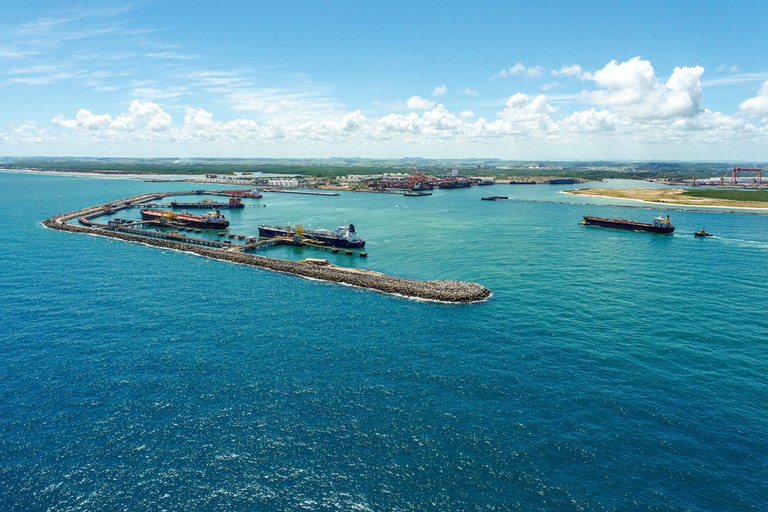 Imagem mostra uma vista aérea do Porto de Suape, localizado no litoral de Pernambuco, Brasil. É possível ver diversos navios atracados, estruturas portuárias ao longo de um quebra-mar e águas azul-turquesa ao redor. Ao fundo, há áreas industriais e vegetação costeira sob um céu claro com algumas nuvens.