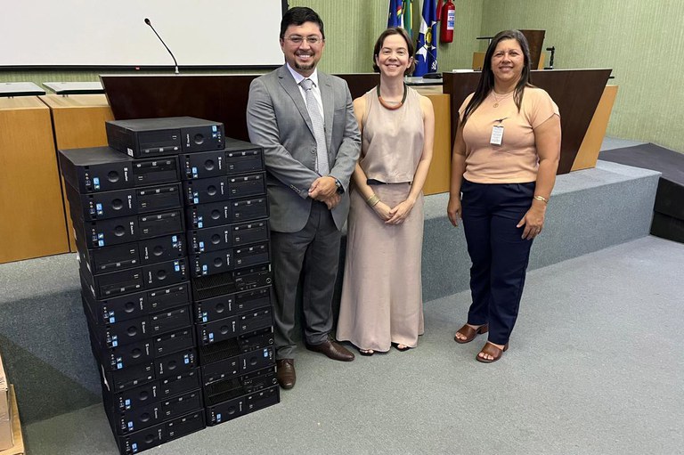 A imagem mostra três pessoas sorrindo em frente a uma pilha de computadores do tipo desktop, empilhados em duas colunas. Eles estão em um auditório, com bandeiras ao fundo e um púlpito, onde ocorreu o momento de doação dos equipamentos.