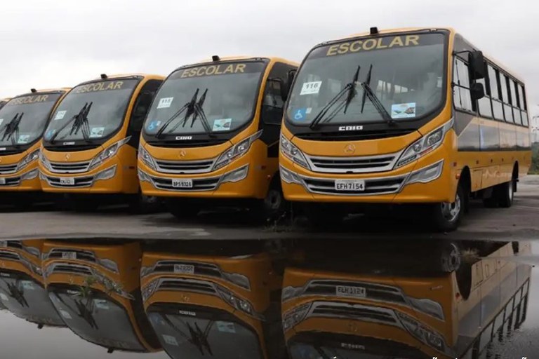 A imagem exibe uma fileira de quatro ônibus escolares amarelos novos, todos com a palavra "ESCOLAR" destacada na parte superior do para-brisa. Os veículos, de modelo micro-ônibus, estão estacionados lado a lado sob um céu nublado, e seus reflexos são vistos em uma poça d'água ou superfície molhada no primeiro plano.