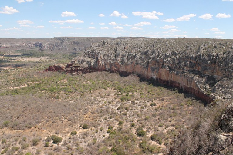Esta imagem captura a grandiosidade do Parque Nacional do Catimbau, em Pernambuco, revelando um vasto vale ladeado por imponentes formações rochosas e chapadas. A vegetação de caatinga, composta por arbustos baixos e áreas de solo exposto em tons de bege e verde seco, estende-se por toda a planície, enquanto os paredões de arenito exibem uma paleta de cores que vai do cinza ao vermelho vibrante, esculpidos pela erosão ao longo de milênios. O horizonte é amplo, sob um céu azul pontuado por nuvens esparsas, transmitindo a atmosfera árida e selvagem característica do sertão nordestino.