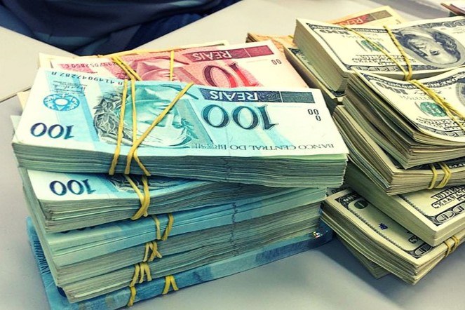 Fotografia de vários maços de dinheiro em cima de uma mesa, alguns de reais e outros de dólares.