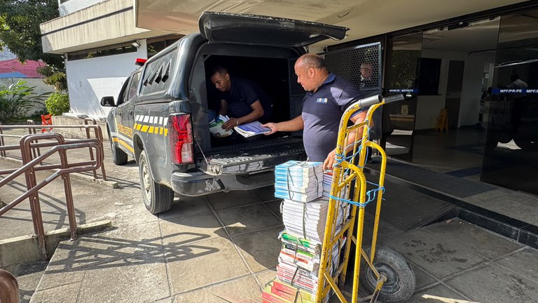 A imagem mostra dois homens colocando livros num veículo oficial do sistema penitenciário. Eles estão transferindo as obras de um carrinho de mão amarelo do MPF para o carro.