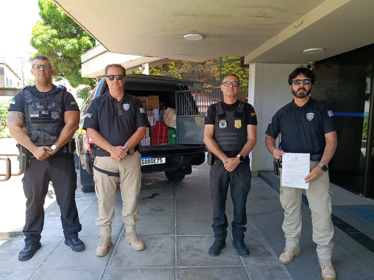 A foto mostra quatro agentes de segurança uniformizados e de óculos de sol alinhados em frente à entrada do MPF em Pernambuco. Atrás deles está um veículo com a mala aberta, em que se encontram empilhados diversos livros e documentos.