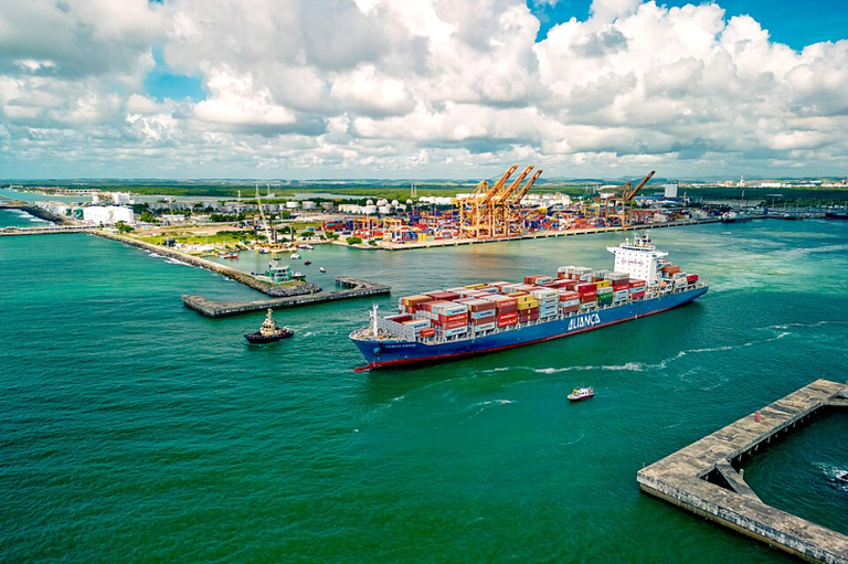 Foto aérea do Complexo Portuário de Suape, mostrando um grande navio de contêineres sendo auxiliado por um rebocador em águas verde-esmeralda, com o terminal e guindastes ao fundo.