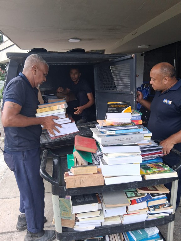 Três homens estão descarregando pilhas de livros do compartimento de carga de um veículo oficial do MPF para um carrinho de transporte de dois andares. Dois deles vestem camisa e calça azuis e organizam os volumes, enquanto um terceiro homem auxilia de dentro do veículo.