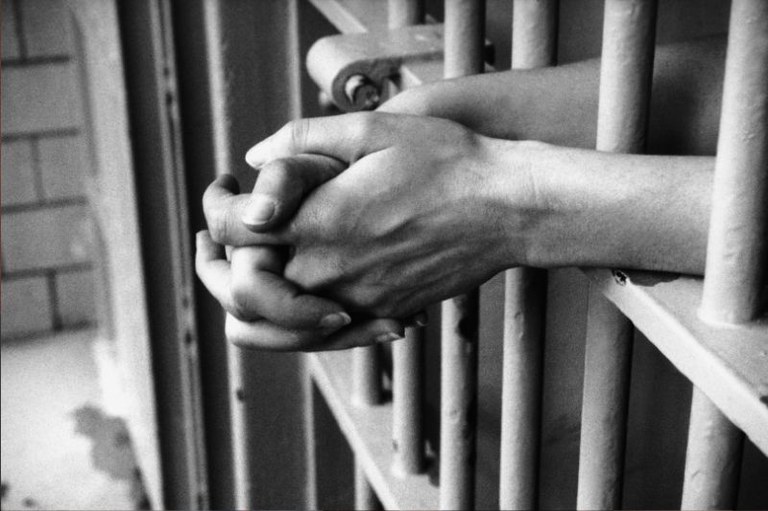 Imagem em preto e branco de uma cela mostrando em detalhe o braço de um preso com as mãos cruzadas para fora