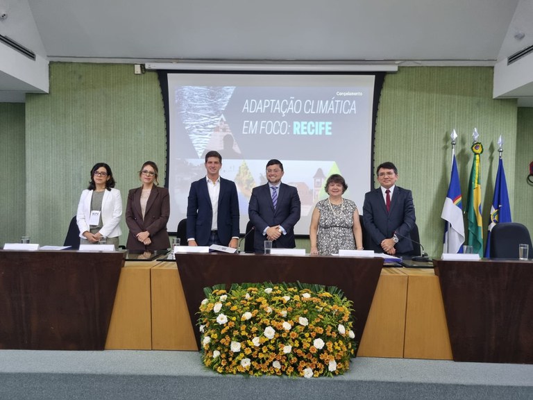 imagem de uma mesa grande com três mulheres e dois homens, vestidos com roupas sociais, atrás o painel do evento Adaptação Climática em Foco: Recife