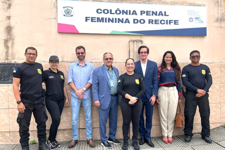 Oito pessoas, entre civis e policiais penais uniformizados, posam para foto em frente à fachada da Colônia Penal Feminina do Recife. O grupo está alinhado horizontalmente diante de uma parede bege com uma placa de identificação da unidade prisional ao topo.