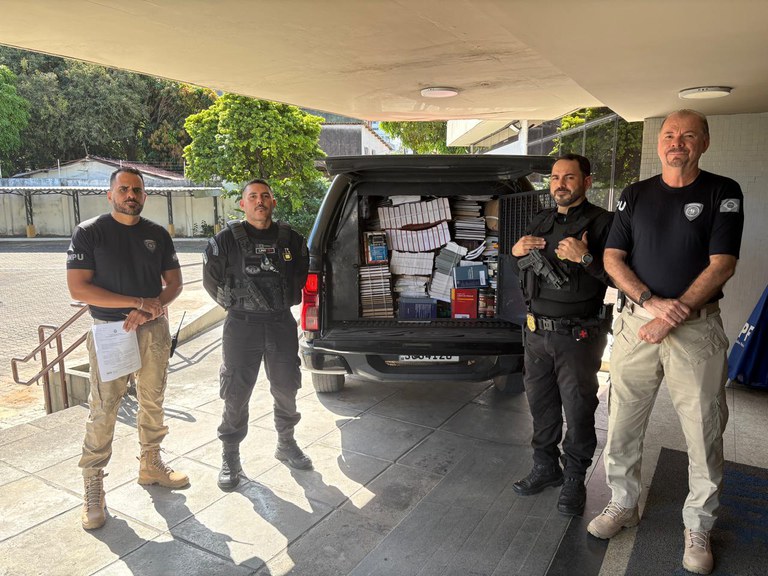 Quatro agentes de segurança uniformizados estão posicionados ao lado da caçamba aberta de um veículo oficial do presídio. A mala está completamente carregada com dezenas de livros e manuais.