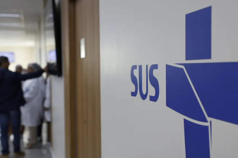 Parede branca em primeiro plano exibindo o logotipo do SUS em azul. Ao fundo desfocado, vê-se o corredor de uma unidade de saúde com pessoas e profissionais vestindo jalecos brancos.