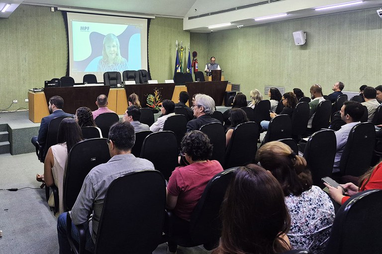 A imagem mostra um auditório com várias pessoas assistindo a uma apresentação. Na frente, dois palestrantes estão posicionados perto de um púlpito, e uma tela exibe uma apresentação com o logotipo "MPF". Bandeiras e um arranjo de flores decoram o ambiente.