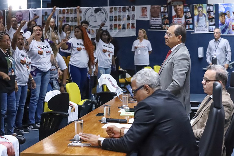 Grupo de pessoas em auditório; mulheres de camiseta branca levantam os punhos em manifestação diante de uma mesa com autoridades sentadas e em pé. Banners e fotos de vítimas estão afixados ao fundo.