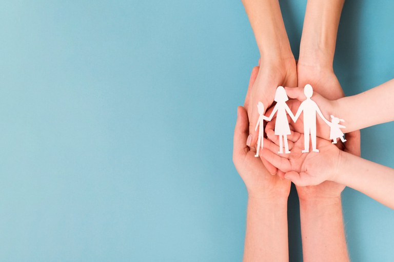 Imagem com fundo azul claro mostra mãos de uma criança por cima das mãos de um adulto. A mão da criança segura um recorte de papel branco com silhueta de uma família composta por pai, mãe e dois filhos.