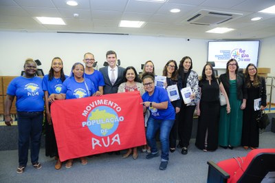 Imagem mostra treze pessoas em pé dentro de auditório. Duas pessoas seguram a bandeira vermelha do Movimento de População de Rua.