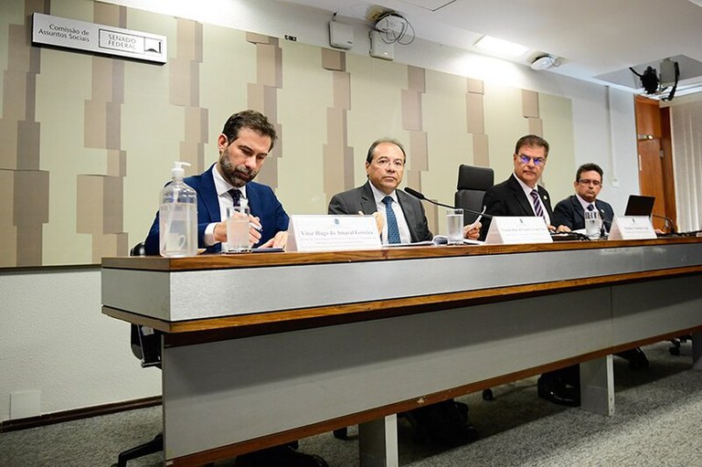 Foto mostra quatro homens sentados atrás de uma bancada, falando com o público.