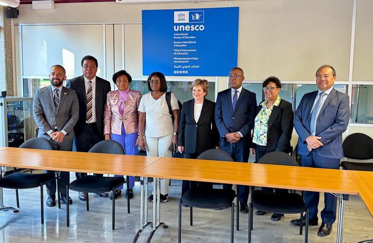 Imagem mostra oito pessoas em pé, com trajes formais, atrás de uma mesa de reuniões.