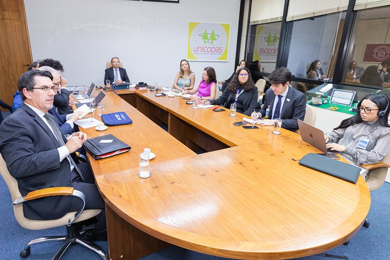 Foto da mesa de reunião com as pessoas sentadas em uma formato de U em uma mesa oval