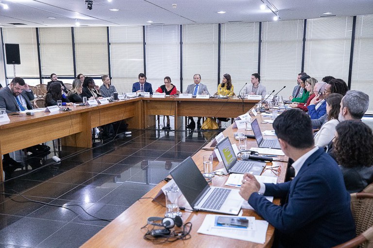Imagem mostra 20 pessoas sentadas em volta de mesa de reunião em formato de U.