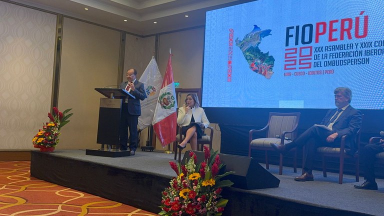 Foto do procurador federal dos Direitos do Cidadão, Nicolao Dino, no XXIX Congresso e Assembleia Geral Ordinária da Federação Iberoamericana de Ombudsperson (FIO). Ele está em pé em um púlpito.   Há outras duas pessoas sentadas no palco.