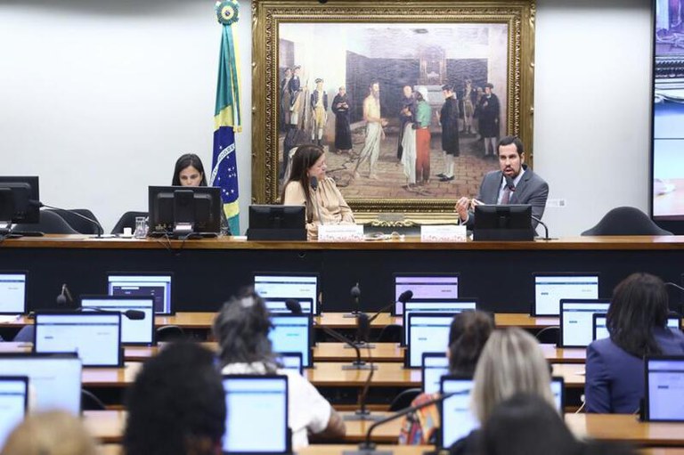 Imagem mostra duas mulheres e um homem sentados atrás de uma bancada. Na audiência, pessoas sentadas à mesa em frente a notebooks.