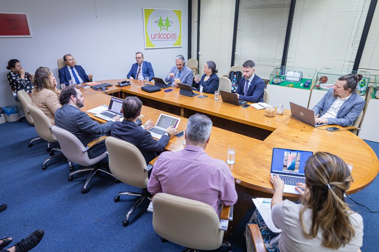 Reunião em sala fechada com cerca de dez pessoas sentadas ao redor de uma mesa oval de madeira, todas usando laptops e materiais de trabalho. O ambiente é corporativo, com janela ampla ao fundo e um painel na parede