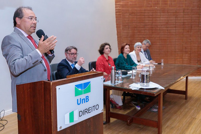 Imagem mostra um homem em pé, atrás do púlpito, fazendo discurso. Atrás dele, uma mesa composta por quatro autoridades.