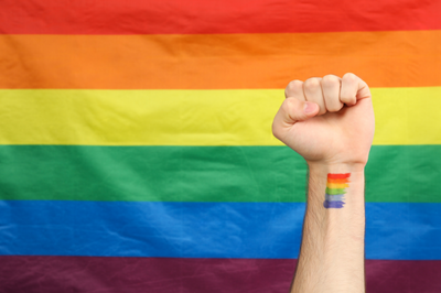 A imagem mostra uma bandeira LGBTQIA+ tem seis cores: vermelho, laranja, amarelo, verde, azul, roxo.Em frente à bandeira, um braço esticado, na vertical, com os punhos cerrados. No braço, uma pequena tatuagem com a bandeira  LGBTQIA+ .