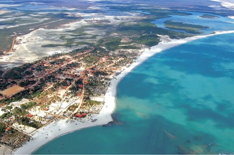 Vista aérea panorâmica de um vilarejo litorâneo, destacando uma extensa faixa de areia branca banhada por mar em tons de azul-esverdeado e estuário ao fundo.