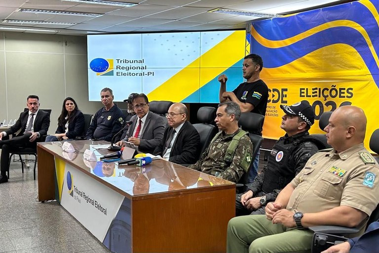 Fotografia horizontal em plano médio que registra uma coletiva de imprensa no Tribunal Regional Eleitoral do Piauí (TRE-PI). Em uma sala bem iluminada, sete homens e uma mulher estão acomodados em fila atrás de uma grande mesa de madeira polida. Entre eles há autoridades civis em trajes formais (como ternos e paletós) e representantes de forças de segurança pública vestindo diferentes fardamentos militares e policiais. Ao centro da mesa, cabos de microfones coloridos de veículos de imprensa estão reunidos em um suporte. À direita, em pé atrás das cadeiras, um intérprete de Libras faz sinais com as mãos. Ao fundo, um telão exibe o logotipo do TRE-PI à esquerda, enquanto um painel amarelo e azul traz as inscrições "Sede Eleições 2026" à direita.