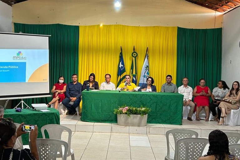 Foto mostra auditório onde foi realizado o evento, ao fundo um cortina verde e amarela, à frente uma mesa com uma tolha verde e 11 pessoas entre homens e mulheres, ao lado esquerdo um telão com  e à frente cadeiras com as pessoas assistindo ao evento