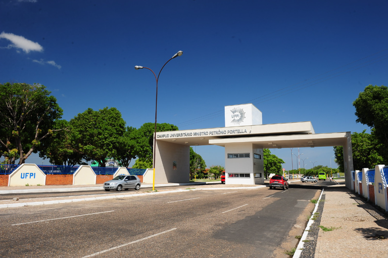 Fotografia mostra o pórtico de entrada da UFPI, com pistas de acesso passando por baixo, num dia de céu azul, sem nuvens. No alto da estrutura, é possível ler "Campus Universitário Ministro Petrônio Portela".