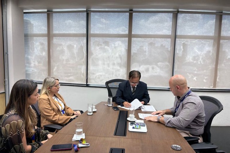 A foto mostra quatro pessoas reunidas em uma sala de reuniões ao redor de uma mesa de madeira, onde um homem de terno escuro assina documentos ao lado de outro homem que manuseia papéis. Duas mulheres sentadas à esquerda observam a formalização do acordo, em uma mesa composta por copos de água, xícaras de café, celulares e crachás, diante de grandes janelas ao fundo