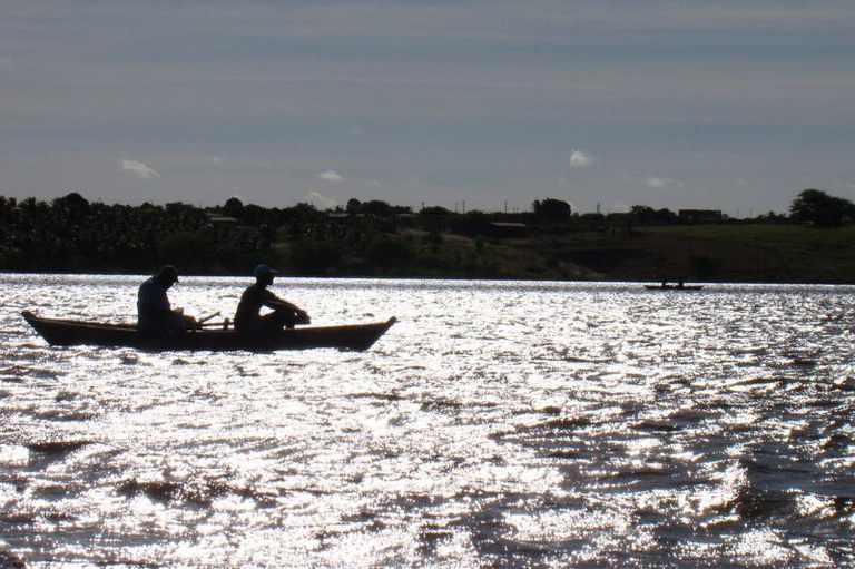 Foto mostra um barco pequeno com dois homens sentados em um rio com o brilho do sol refletindo na água