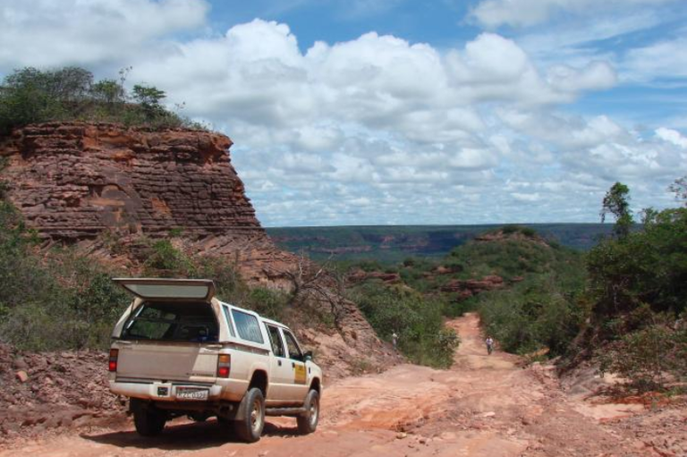 Foto mostra caminhonete branca em estrada de terra avermelhada com a caçamba aberta, em uma paisagem de cerrado com formações rochosas íngremes.
