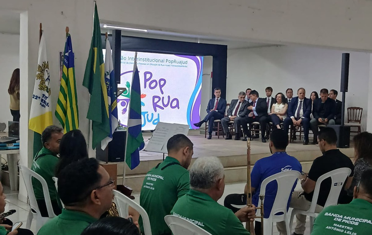 Imagem do evento institucional PopRuaJud em Picos (PI, ocorrido no dia 28 de novembro de 2025. No ambiente, há várias pessoas sentadas nas cadeiras brancas, de costas para a câmera, ou parcialmente visíveis. Muitos deles estão vestindo camisas ou camisetas verdes que parecem ser uniformes. No fundo, sobre um palco, há um grupo de autoridades ou convidados sentados em cadeiras estofadas escuras. Todos estão vestindo trajes sociais, como ternos. Há cerca de dez pessoas visíveis nesta área.