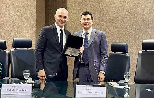 Fotografia que registra entrega de placa em homenagem ao procurador regional da República Marco Aurélio Adão pelo procurador-chefe da Procuradoria da República no Piauí, Marco Túlio Caminha. Os dois estão atrás de uma mesa de conferência em um ambiente institucional. O foco central é a entrega de uma placa de homenagem (dentro de um estojo de veludo preto aberto). À frente de cada um, há placas de identificação com o logotipo do MPF e seus respectivos nomes.