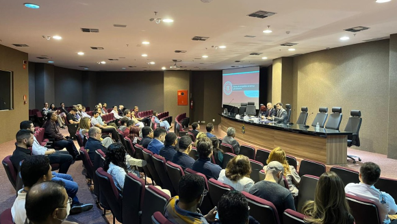 A imagem retrata autoridades, o palestrante e participantes durante o primeiro Ciclo de Palestras Temas Jurídicos Contemporâneos, no auditório da Procuradoria da República no Piauí, no dia 4 de abril de 2025. Na bancada, há três pessoas, dois homens e uma mulher. Na plateia, os participantes estão sentados. Ao lado da bancada há um telão com a imagem inicial da palestra e um púlpito.