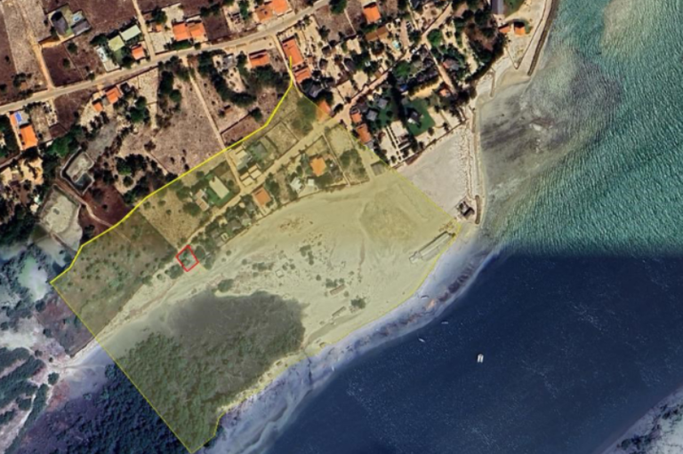 Vista aérea de satélite de uma área costeira urbana com rio e praia, onde uma porção de terra próxima à água está delimitada por um contorno amarelo.