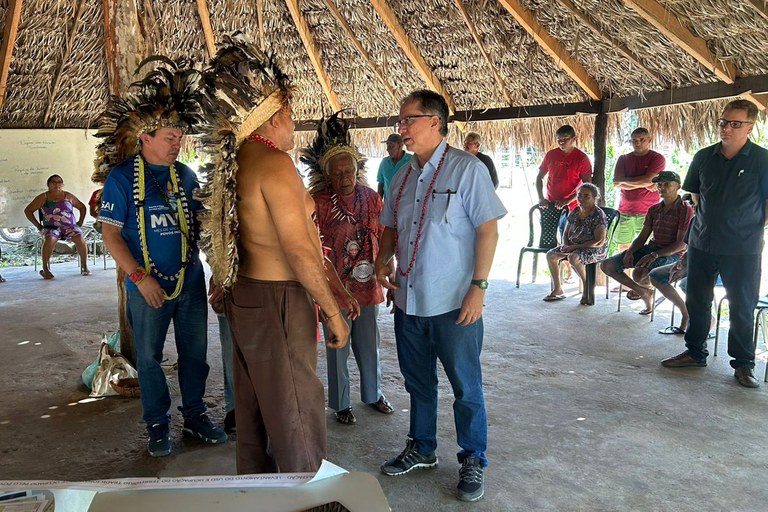 Descrição da imagem: Fotografia de visita técnica realizada pelo Ministério Público Federal na comunidade indígena Tabajara, no município de Piripiri, no Piauí. Ao centro, destacam-se lideranças indígenas utilizando cocares de penas imponentes e colares tradicionais. Um deles, de costas para a câmera, usa um cocar longo que desce pelas costas, enquanto outro, de frente, veste uma camiseta azul com a inscrição "Mês de Vacinação dos Povos Indígenas". À direita do centro, um homem de pele clara, óculos e camisa social azul clara (também usando um colar indígena) conversa diretamente com as lideranças. Sua postura indica um diálogo atento e respeitoso. O encontro ocorre em um espaço amplo e aberto, com um telhado rústico de palha sustentado por vigas de madeira. Ao fundo, vê-se um quadro branco com anotações manuscritas, possivelmente os tópicos da reunião. Diversas pessoas da comunidade e outros assessores estão sentados em cadeiras de plástico ao redor do espaço, observando a interação central. O grupo é diversificado, incluindo homens, mulheres e jovens.