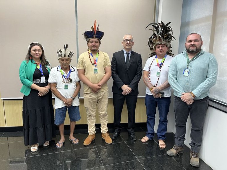 Registro de reunião do procurador da República Marco Aurélio Adão com equipe representantes de comunidades indígenas e Funai. Na imagem, cinco homens e uma mulher estão em uma sala de reunião, em pé. O procurador ladeado por dois grupos, três homens e uma mulher de um lado e dois homens do outro. Os indígenas usam adorno na cabeça.