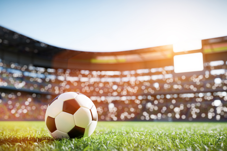 A imagem destaca uma bola de futebol clássica em primeiro plano, posicionada sobre o gramado verde e bem cuidado de um grande estádio. Ao fundo, as arquibancadas repletas de espectadores aparecem completamente desfocadas através de um efeito bokeh, sob a luz intensa do sol que ilumina o topo da arena. Essa composição visual transmite com perfeição a atmosfera vibrante e a expectativa que antecedem um grande dia de jogo.