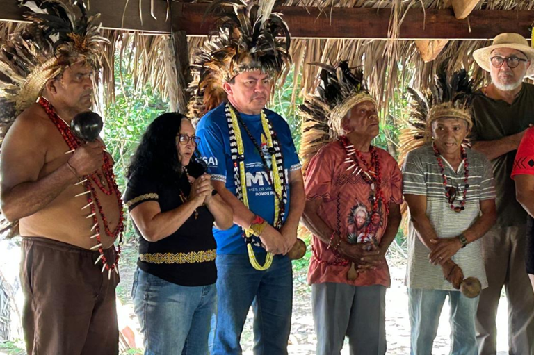 Grupo de pessoas reunidas sob uma estrutura com teto de palha, com uma mulher e cinco homens indígenas usando cocares de penas, colares tradicionais e segurando maracás.