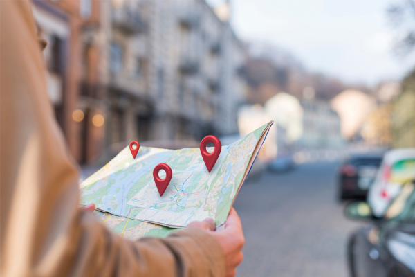 Pessoa segura um mapa em uma rua urbana, com marcadores de localização destacados em pontos específicos. A imagem remete a uma busca por destinos ou planejamento de rota.