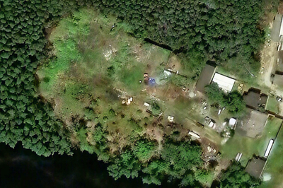  imagem aérea de uma área às margens de um corpo d’água escuro, cercada por vegetação densa; no centro, há uma clareira que foi desmatada.