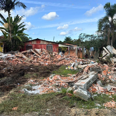 foto mostra grande quantidade de entulho, resultante da demolição de construção irregular; ao fundo, aparecem casas simples, vegetação e céu azul com nuvens dispersas.