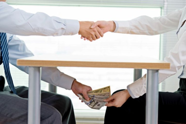 foto colorida de duas pessoas em trajes sociais, sem mostrar os rostos; elas apertam as mãos sobre uma mesa enquanto, por baixo dela, uma delas entrega secretamente um maço de notas de dinheiro para a outra.