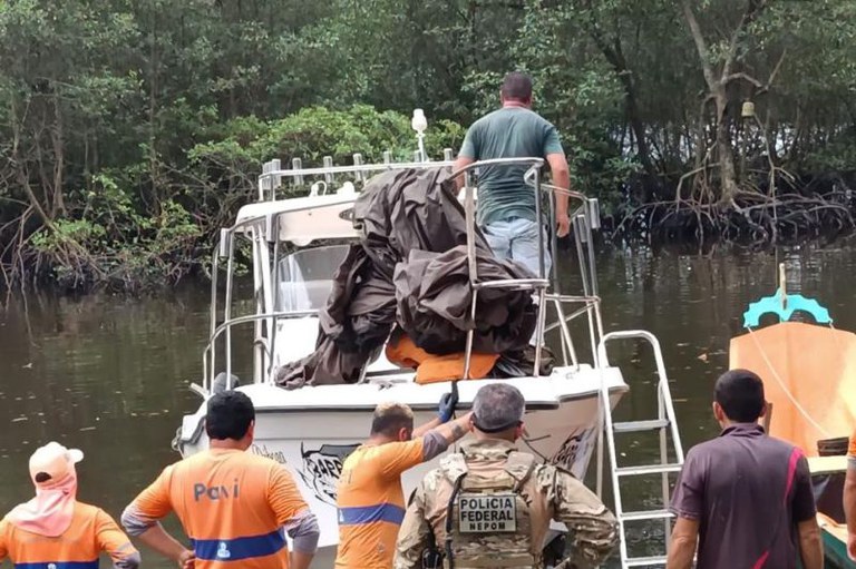 Foto mostra uma embarcação no rio com um homem dentro, o mangue ao fundo e na orla cinco homens
