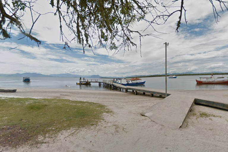 Vista de um trapiche de concreto que se estende de uma praia de areia clara em direção a águas calmas. Várias embarcações pequenas e coloridas estão atracadas ao longo da estrutura ou ancoradas próximas à costa. Algumas pessoas são vistas sobre o trapiche. A cena é emoldurada por galhos de árvores em primeiro plano e, ao fundo, montanhas azuis sob um céu nublado com aberturas de azul.